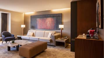 A modern hotel room with a sofa, armchairs, a coffee table, a side table, and a large abstract painting on the wall.