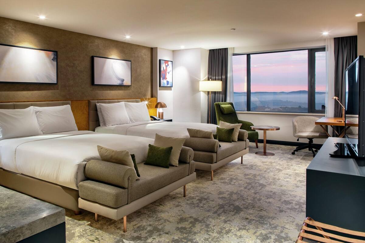A hotel room with two beds, a desk, a chair, and a view of the cityscape.