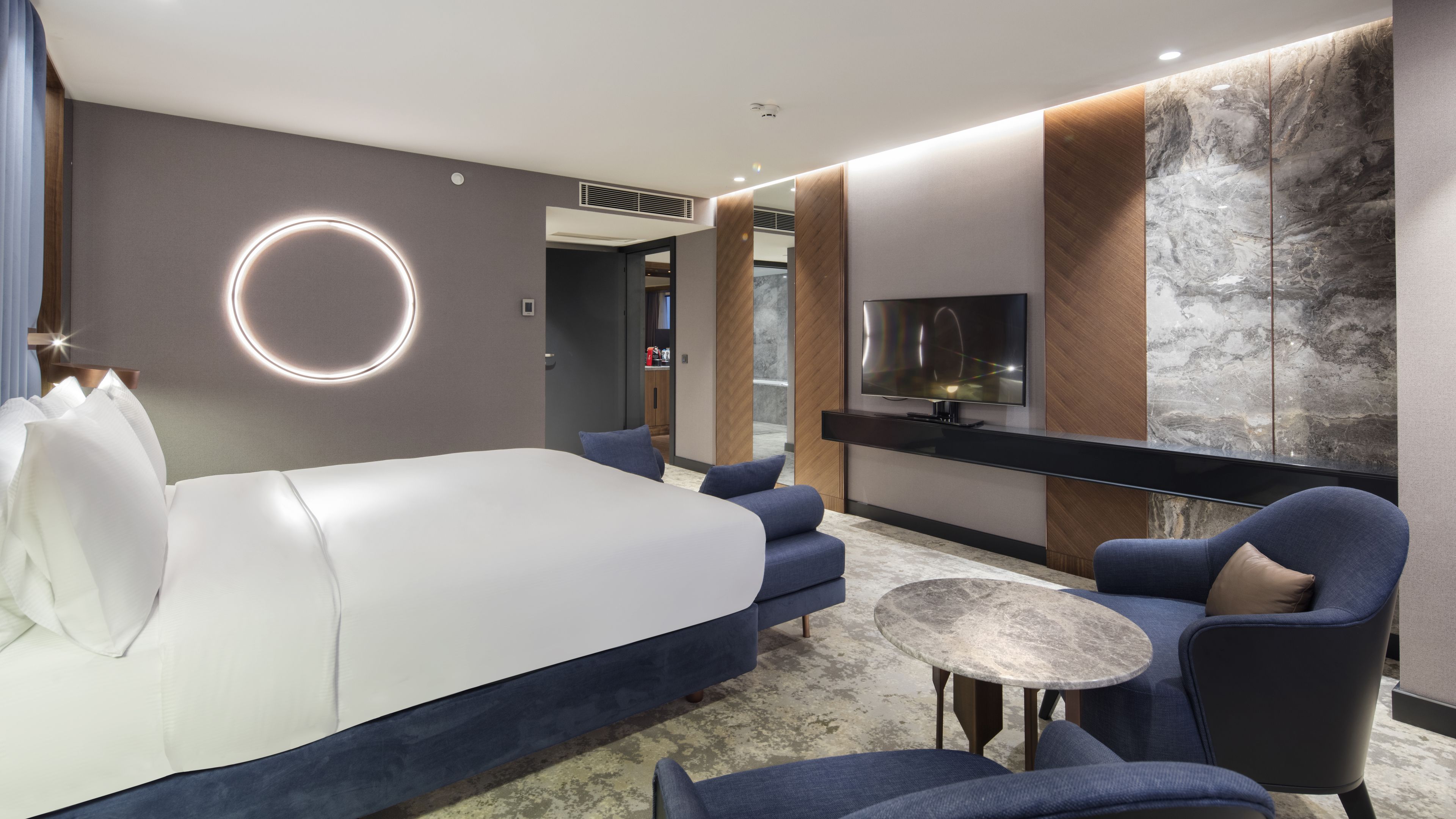 A modern hotel room with a large bed, a TV, a round mirror, and a seating area with a small table.