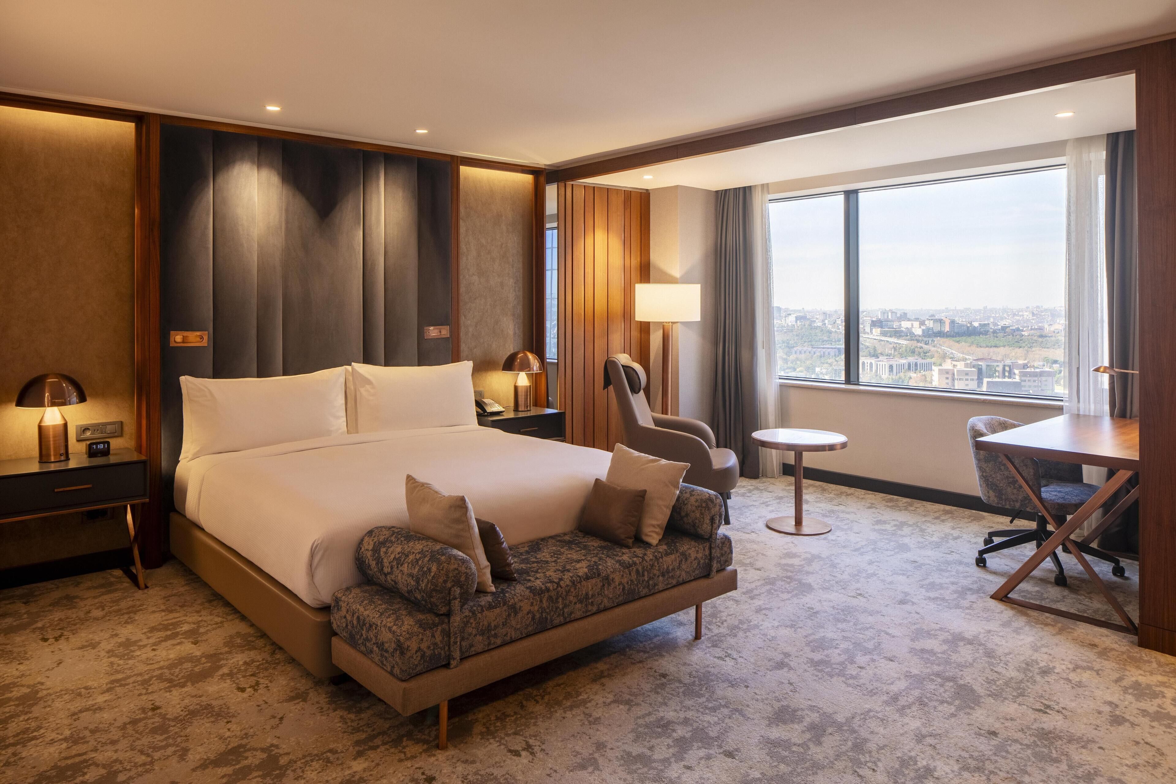 A hotel room with a large bed, a sofa, a desk, and a view of the city.