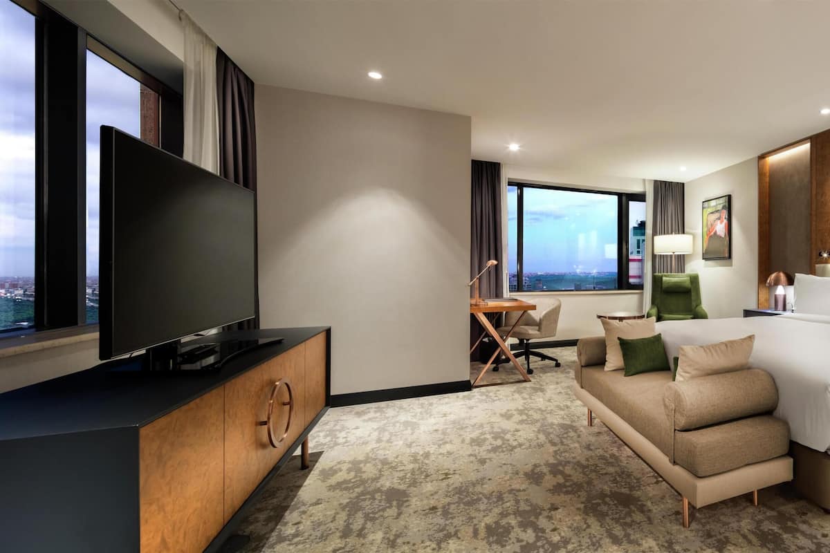 A modern hotel room with a large bed, a TV, a desk, and a view of the city.