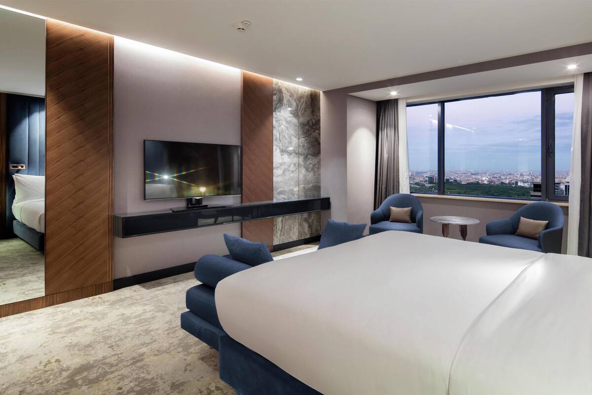 A modern hotel room with a large bed, a TV, a seating area with chairs, and a city view.