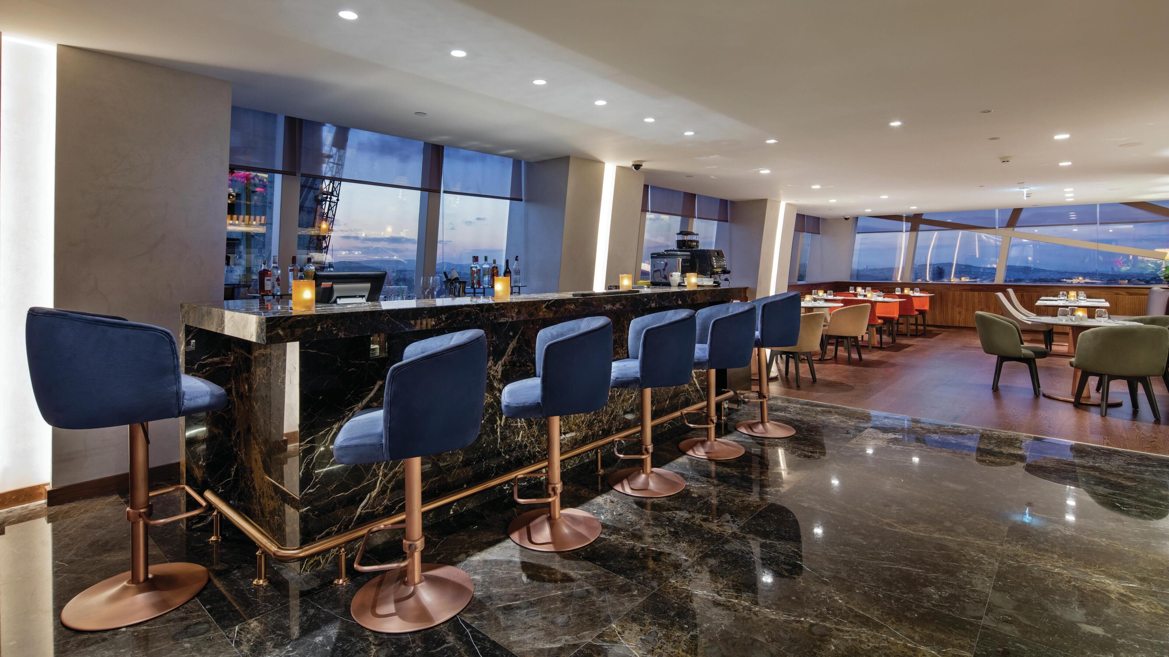 Zona de bar con mostrador de mármol, varios taburetes y vistas panorámicas de la ciudad a través de amplios ventanales.