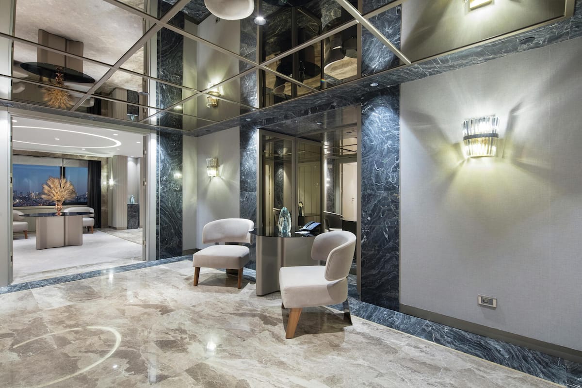 A modern lobby with marble floors, two upholstered chairs, a reception desk, and a large mirror reflecting the interior.