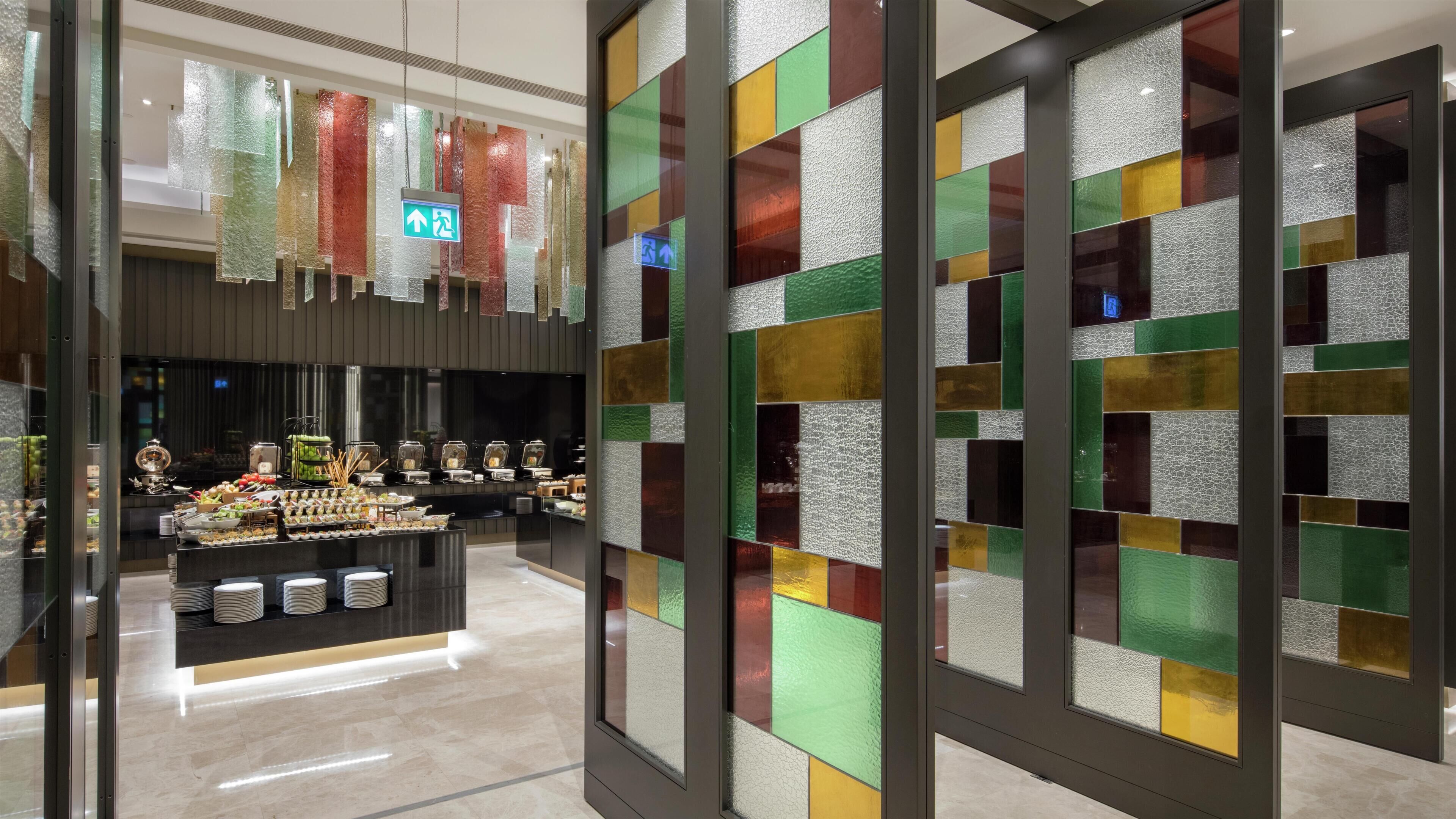 A hotel lobby with a reception desk, a buffet area, and a wall with colorful glass panels.
