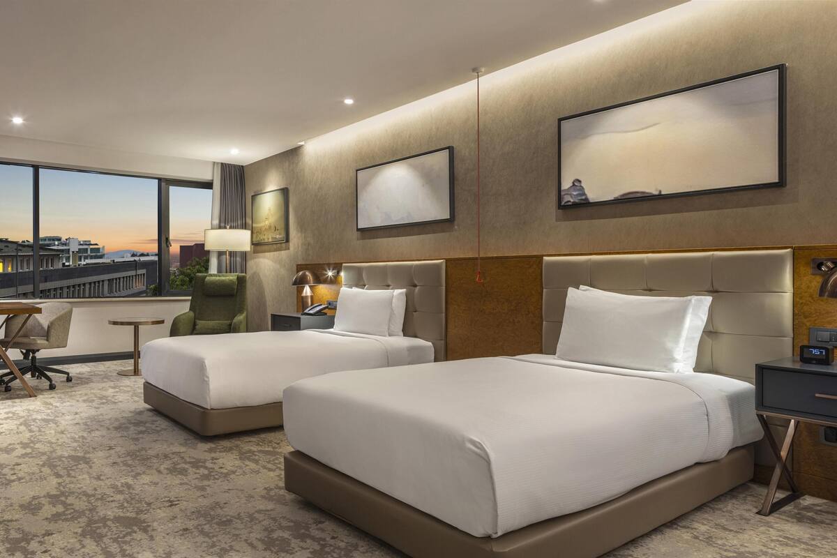 A hotel room with two beds, a desk, a chair, a nightstand, and a large window with a city view.