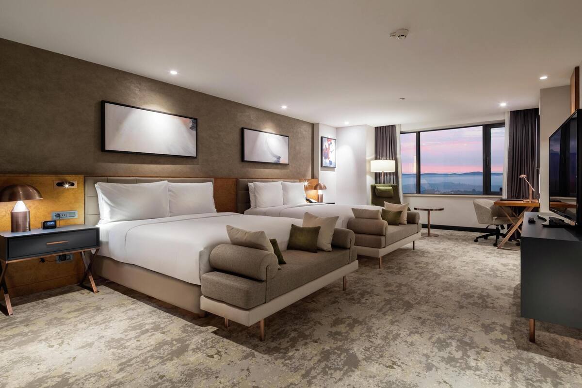 A modern hotel room with a large bed, two bedside tables, a desk, a chair, and a view of the cityscape.