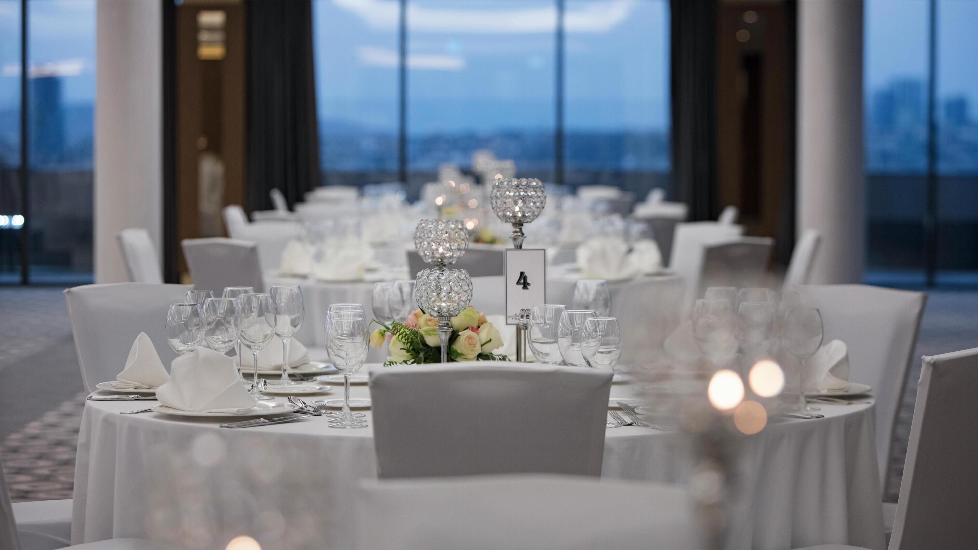A banquet hall with round tables, each with a number sign, white tablecloths, and clear glassware.