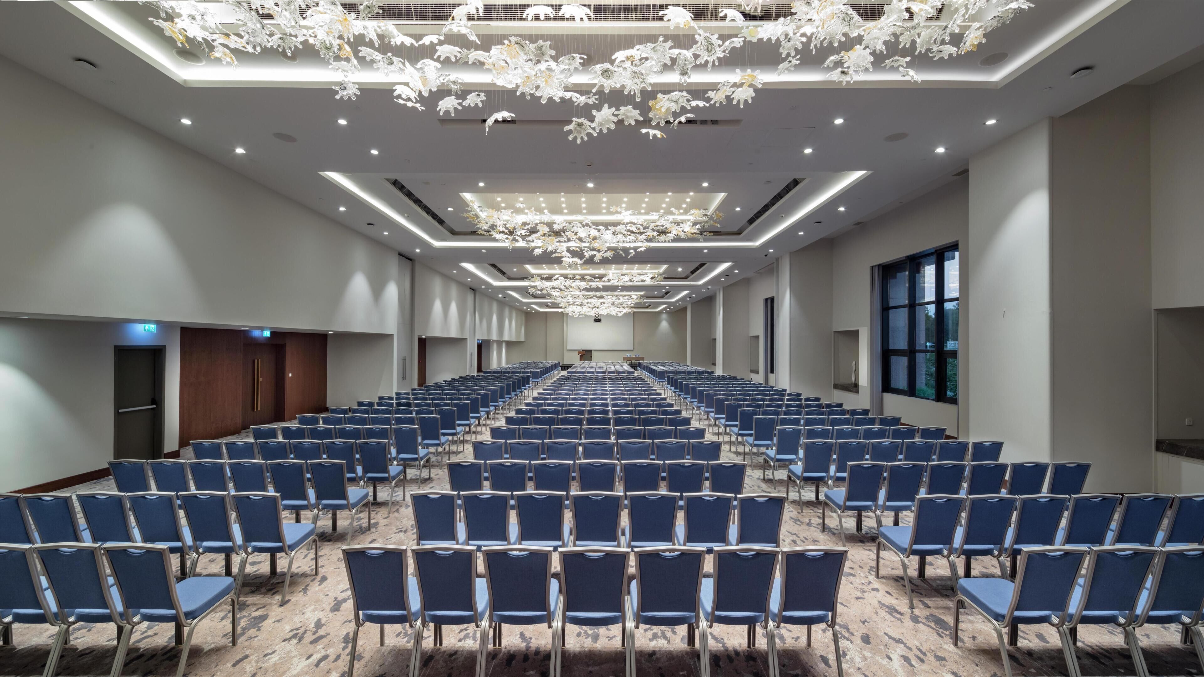 A conference room with rows of blue chairs, a large screen at the front, and a modern chandelier.