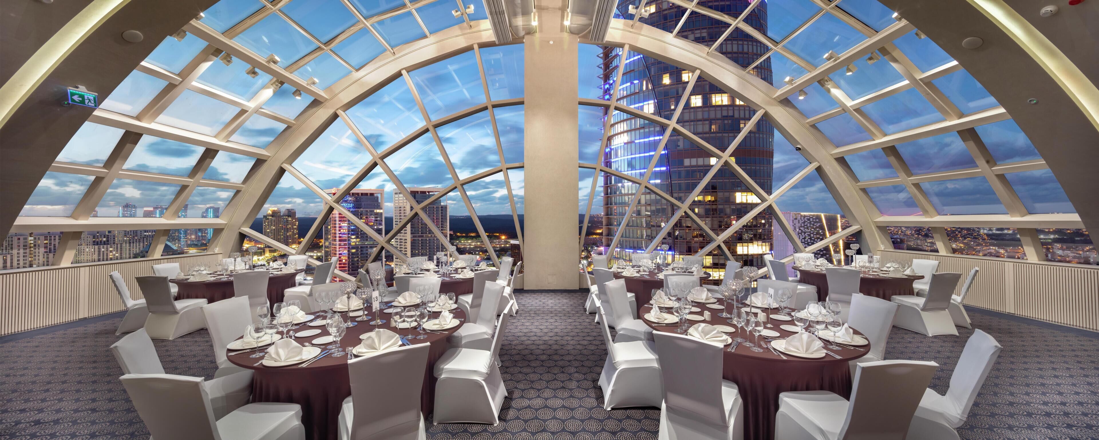 A dining area with tables set for service, a city view through a glass ceiling, and a modern architectural design.