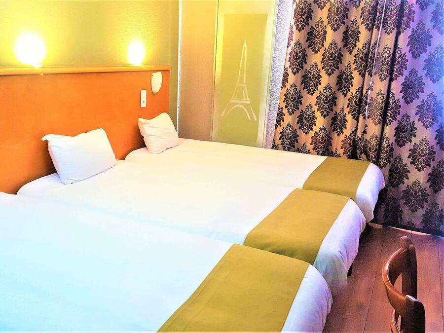 A hotel room with a bed, two pillows, a nightstand, a chair, and a curtain with a floral pattern.