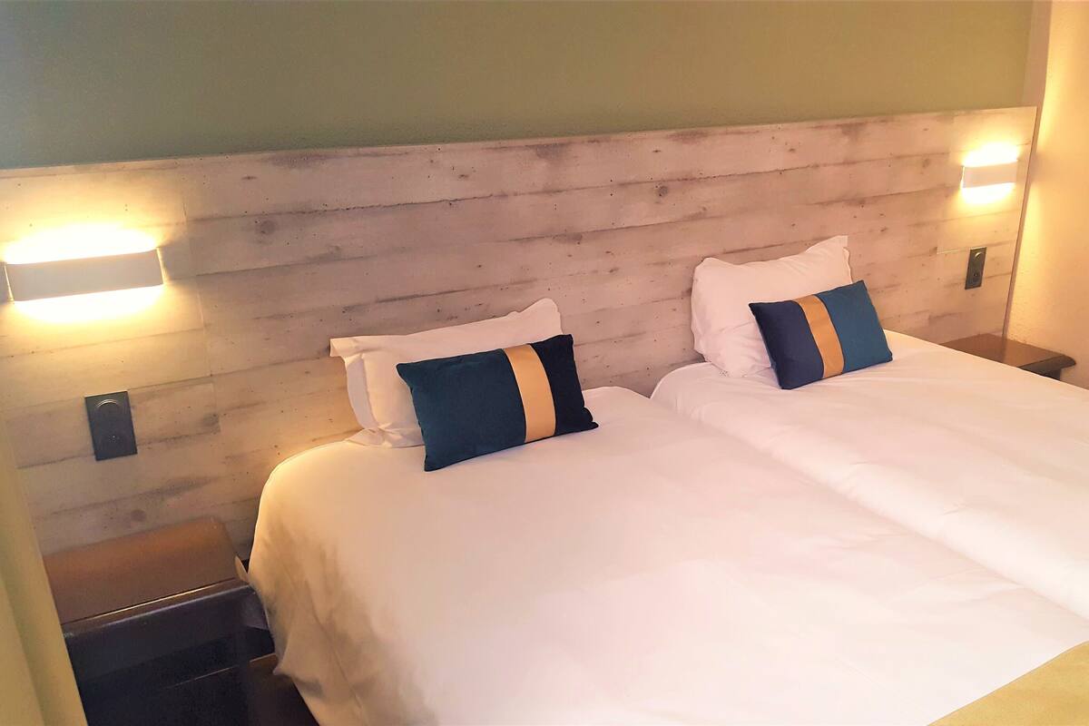 A double bed with white bedding and two pillows, one with a blue and gold stripe. There are two bedside tables with lamps on either side of the bed.