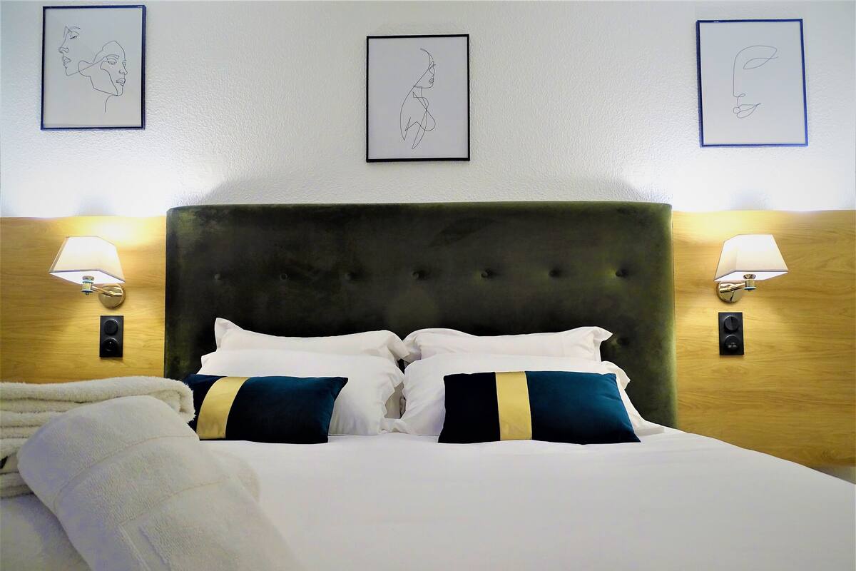 A neatly made bed with white and blue pillows, a white blanket, and a white towel. There are two wall-mounted lamps on either side of the bed.