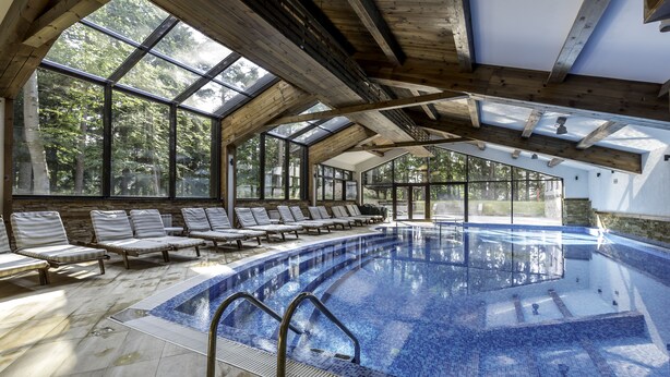 Une piscine intérieure avec des poutres en bois, de grandes fenêtres et des chaises longues baignées de lumière.