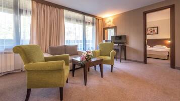 Ein Hotelzimmer mit einer Couch, zwei Sesseln, einem Couchtisch mit Blumen, einem Fernseher und einem Bett.