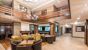 Un hall d’entrée spacieux avec un plafond haut, un espace de détente central avec des tables et des chaises, une cheminée et un bar doté d’un grand écran.