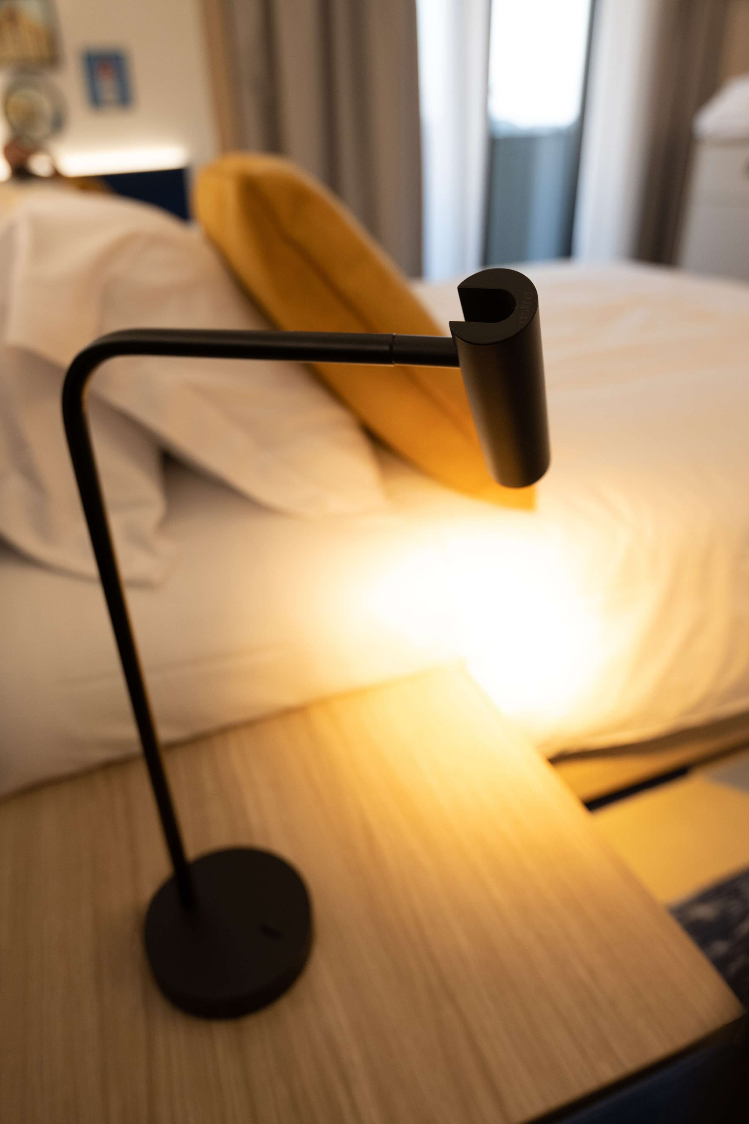 Una mesita de noche con una lámpara moderna, una cama con ropa blanca y una almohada amarilla.