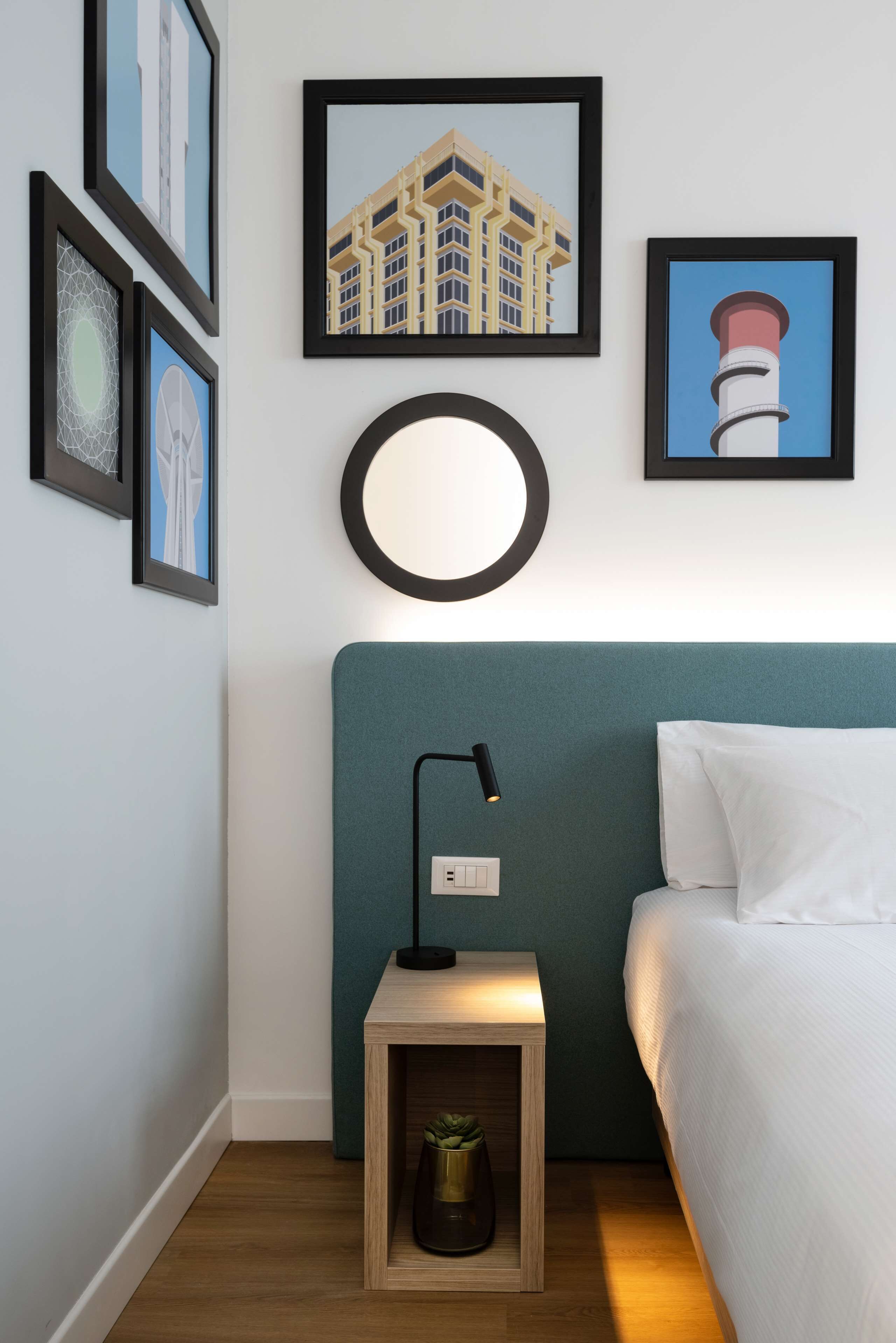 Une chambre d’hôtel avec une tête de lit de couleur turquoise, une table de chevet avec une lampe, une petite plante et des tableaux encadrés accrochés au mur.