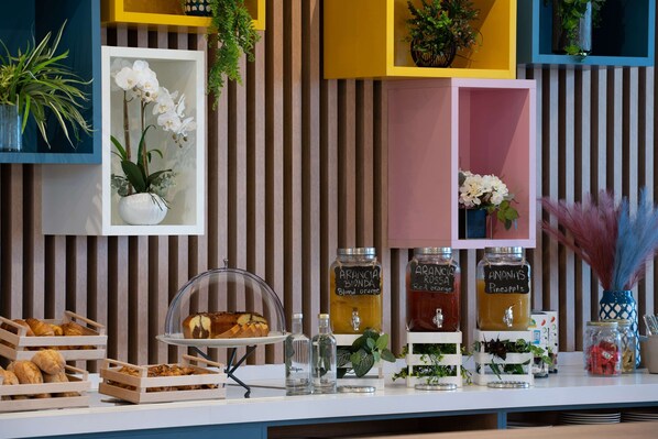 A breakfast buffet with various pastries, a fruit display, and beverage dispensers labeled with different flavors.