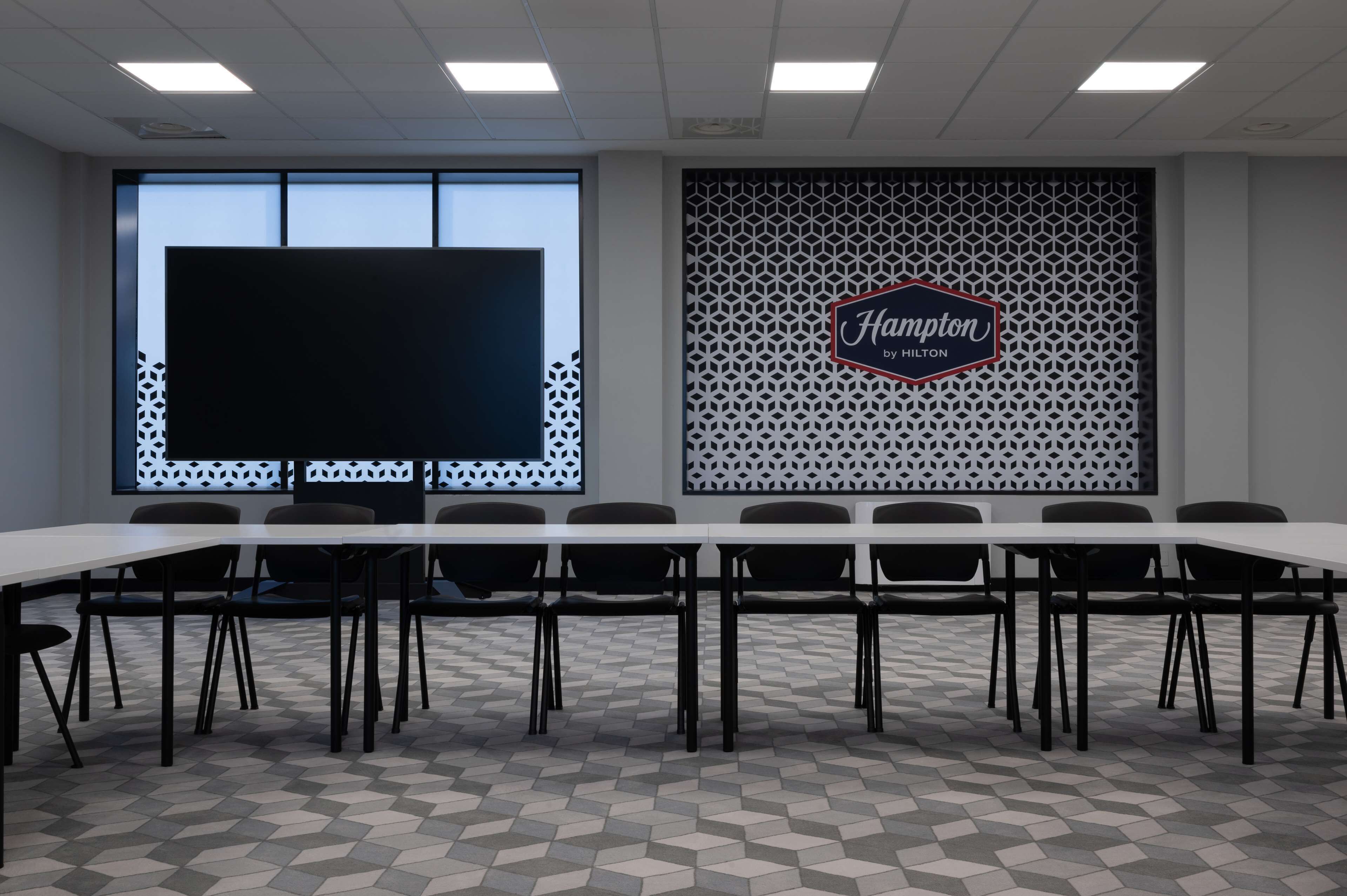 A conference room with a long table, chairs, a large screen, and a decorative wall with a 'Hampton by Hilton' sign.