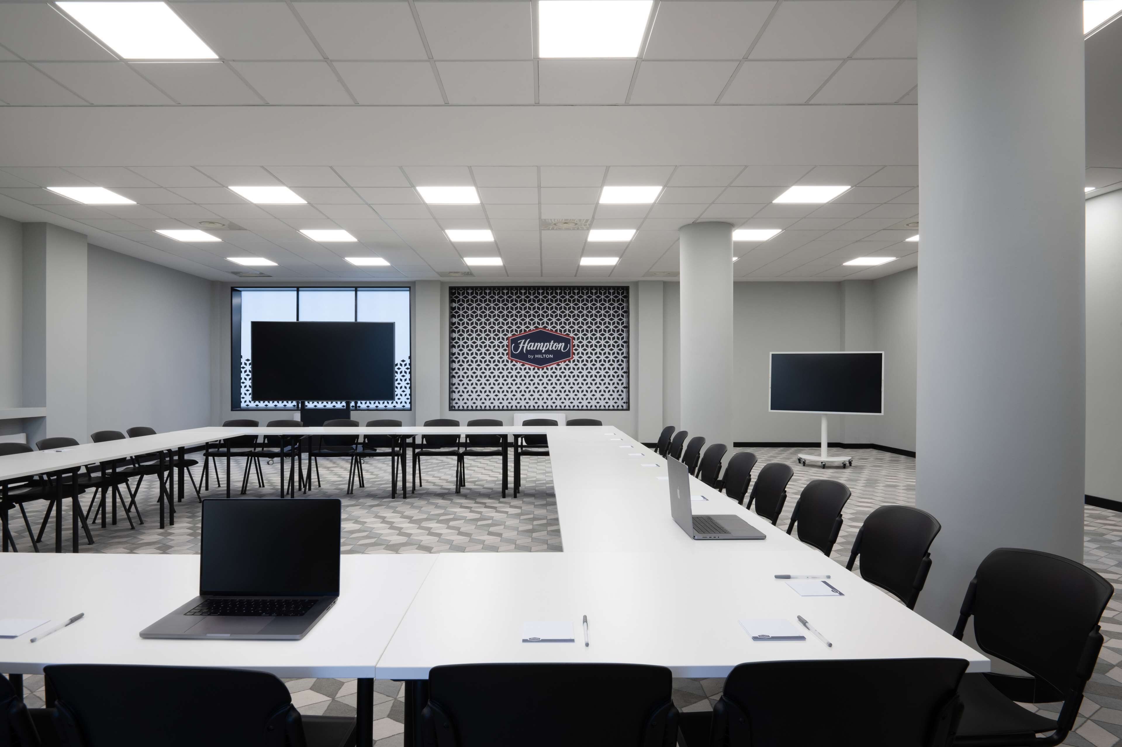 A conference room with a long table, chairs, a laptop, a projector screen, and a Hampton by Hilton sign.