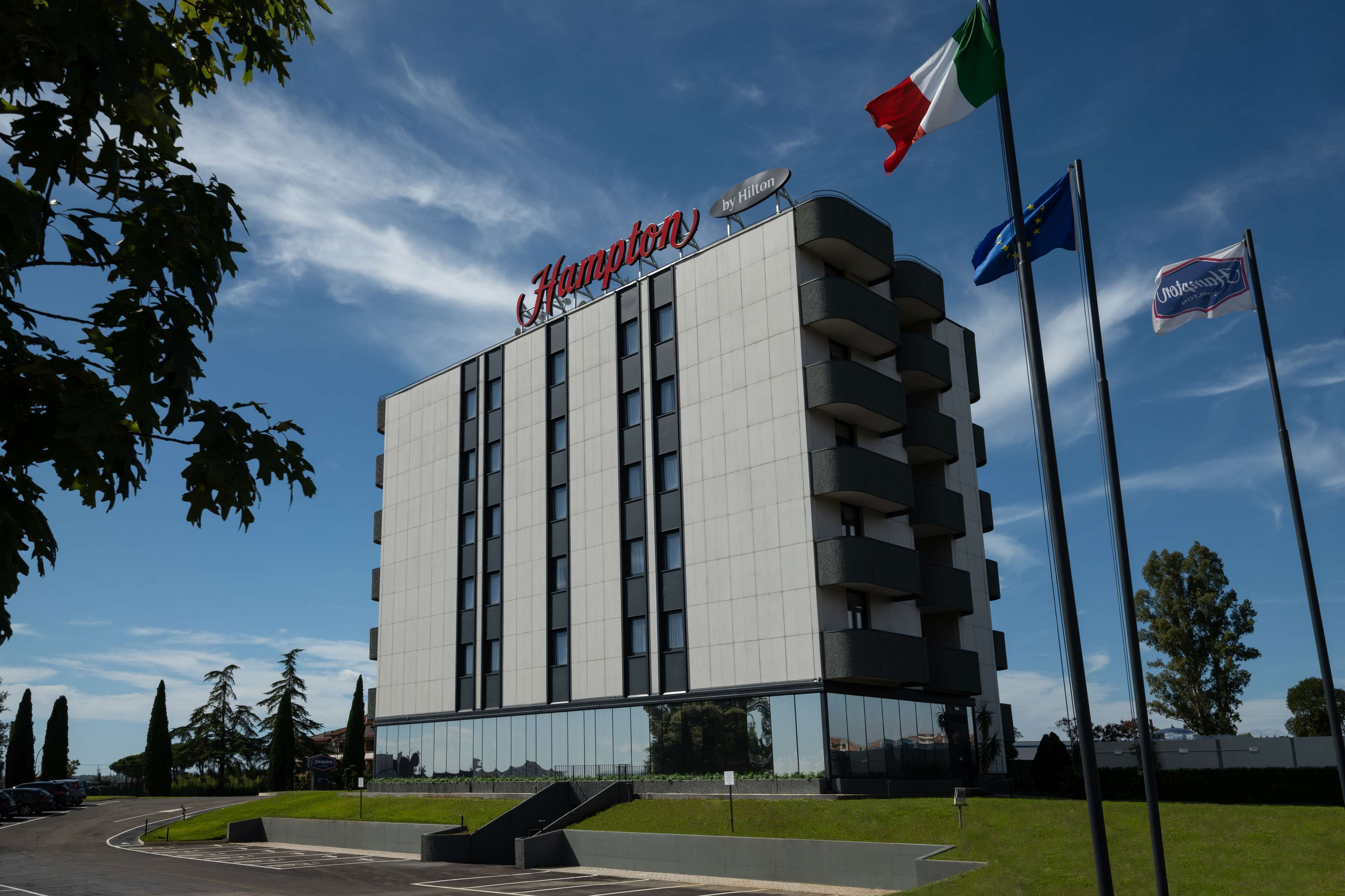 Un moderno edificio de hotel con un llamativo letrero de 'Hampton', varias banderas y un cielo despejado.