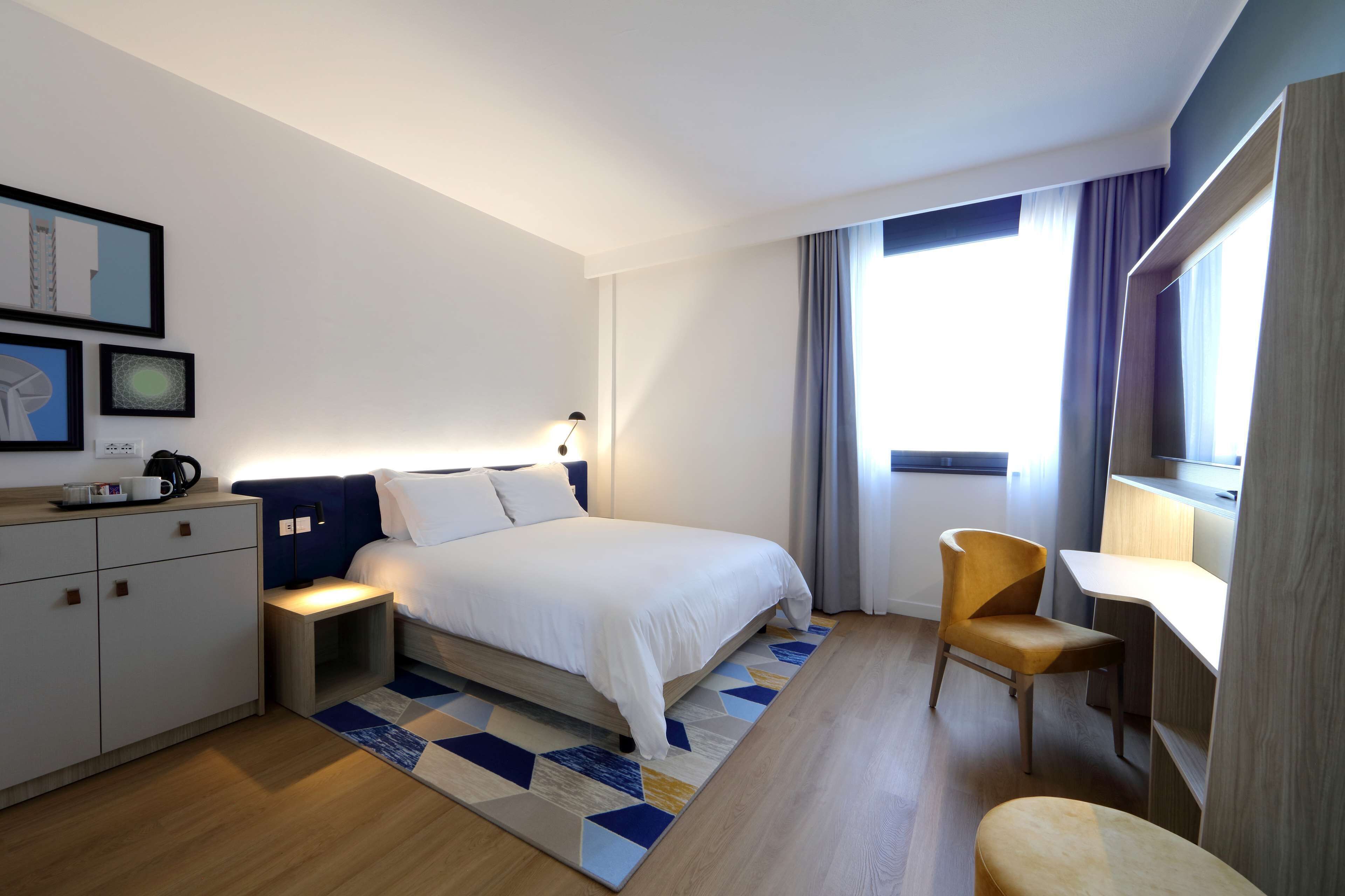 Habitación de hotel moderna con una cama bien tendida, un escritorio y una silla. Hay una mesita de noche con lámpara, un armario y una ventana con cortinas.