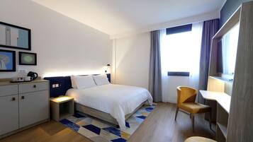 Habitación de hotel moderna con una cama bien tendida, un escritorio y una silla. Hay una mesita de noche con lámpara, un armario y una ventana con cortinas.