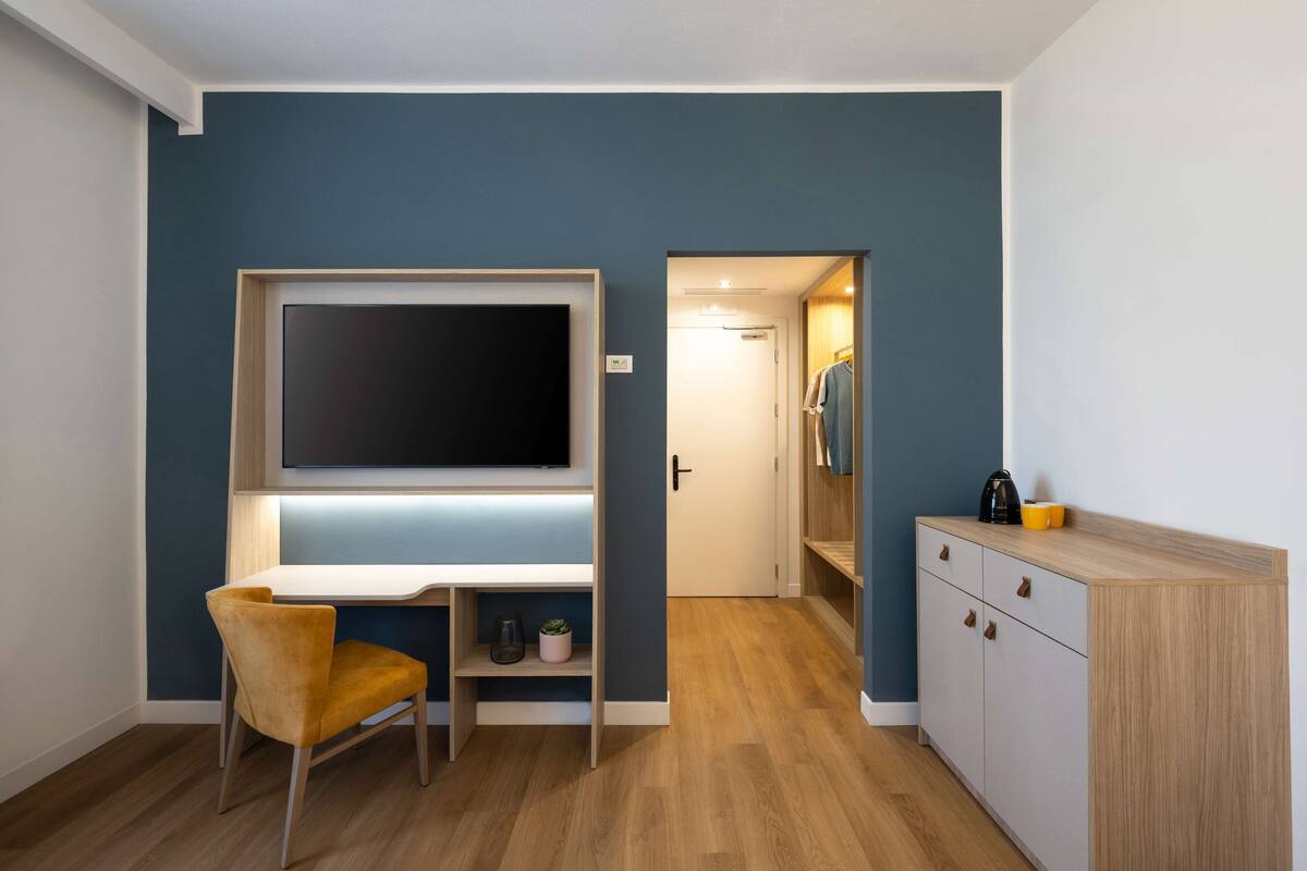 A modern room with a large flat-screen TV mounted on a wall, a desk with a chair, a small cabinet, and a door leading to a bathroom.