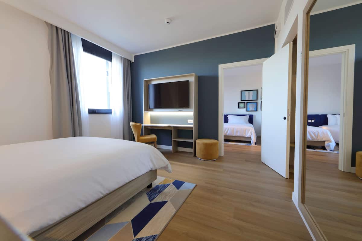 A modern hotel room with a large bed, a desk with a flat-screen TV, and a second bed in the background.