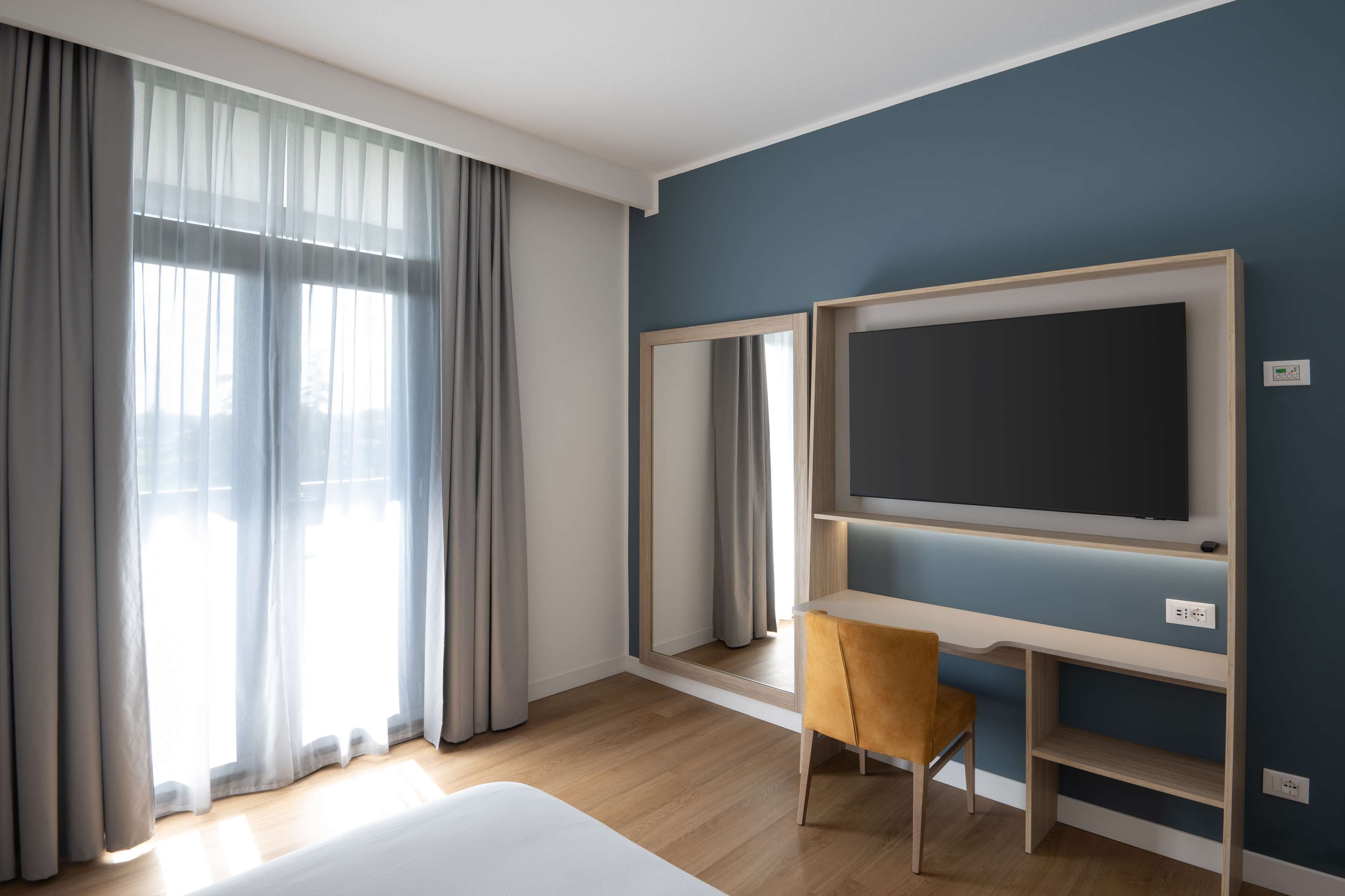 Una camera d'albergo con un letto, una televisione a parete, una sedia e un'ampia finestra con tende.