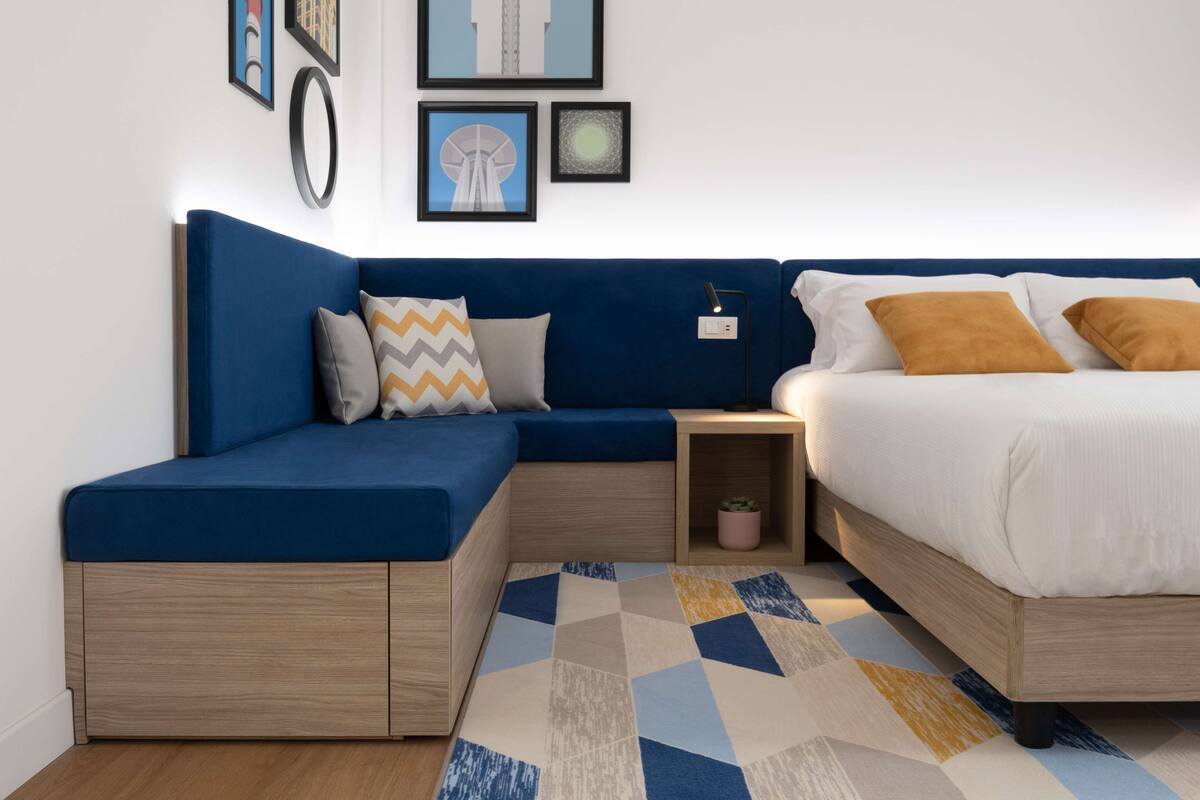 A modern bedroom with a blue sofa bed, a separate bed with white bedding, and a geometric patterned rug.