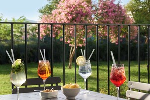 Una mesa con cuatro bebidas y un bol de nueces, frente a un jardín con flores rosadas y una barandilla metálica.