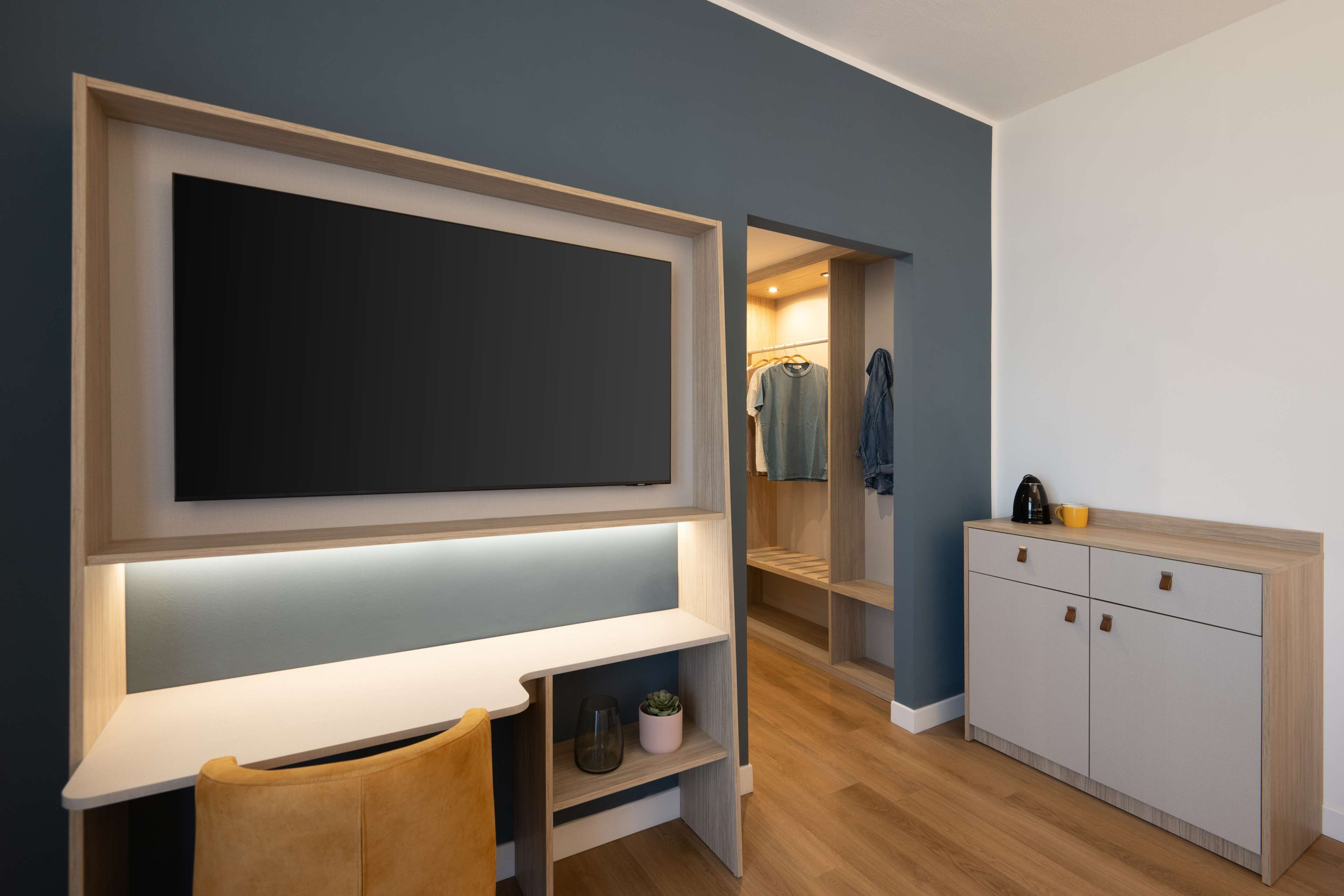 A modern room with a large flat-screen TV mounted on a wall, a wooden desk, a chair, a wardrobe, and a small cabinet with drawers.