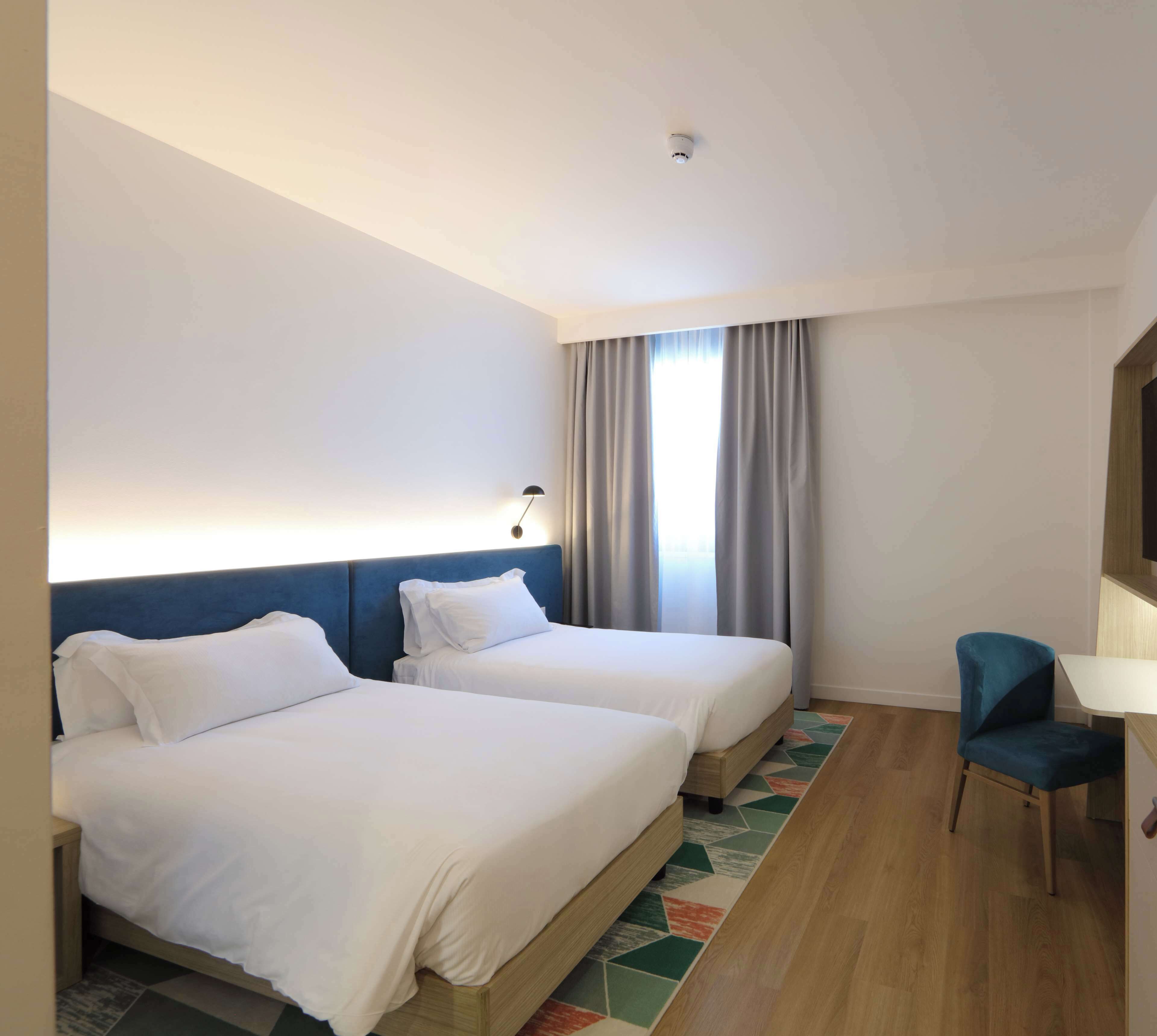 Ein Hotelzimmer mit zwei Betten, einem Schreibtisch, einem Stuhl und einem großen Fenster mit Vorhängen.
