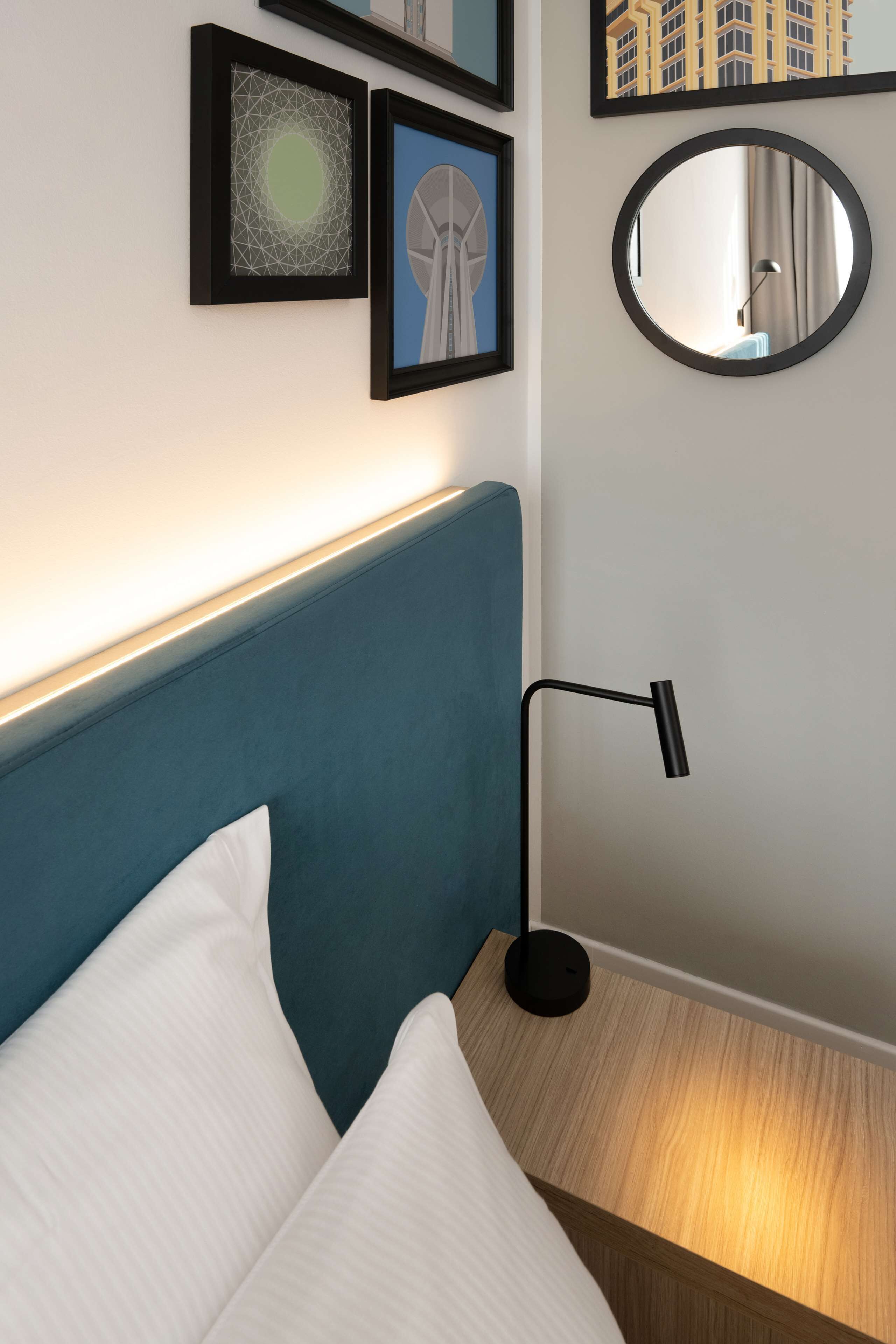 A neatly made bed with white pillows, a blue headboard, and a bedside table with a lamp. There are framed pictures on the wall above the bed.