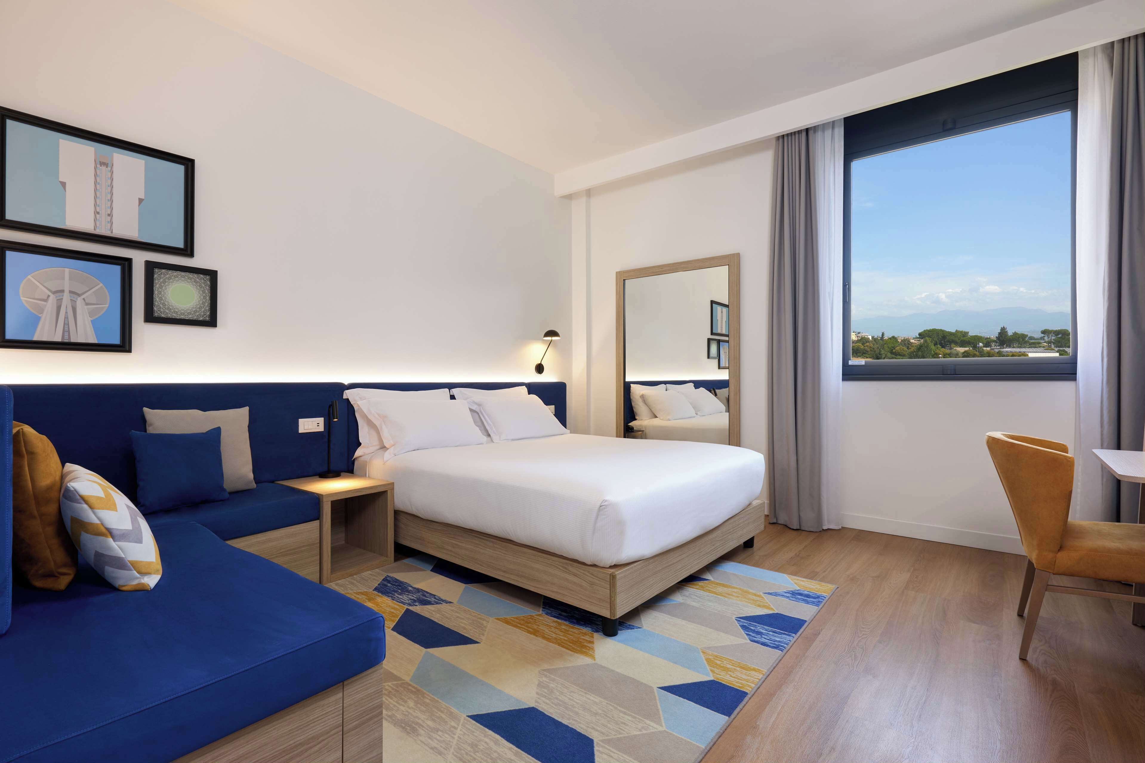 Habitación de hotel moderna con una cama grande, un sofá azul, una mesa de comedor y un ventanal con vistas a la ciudad.