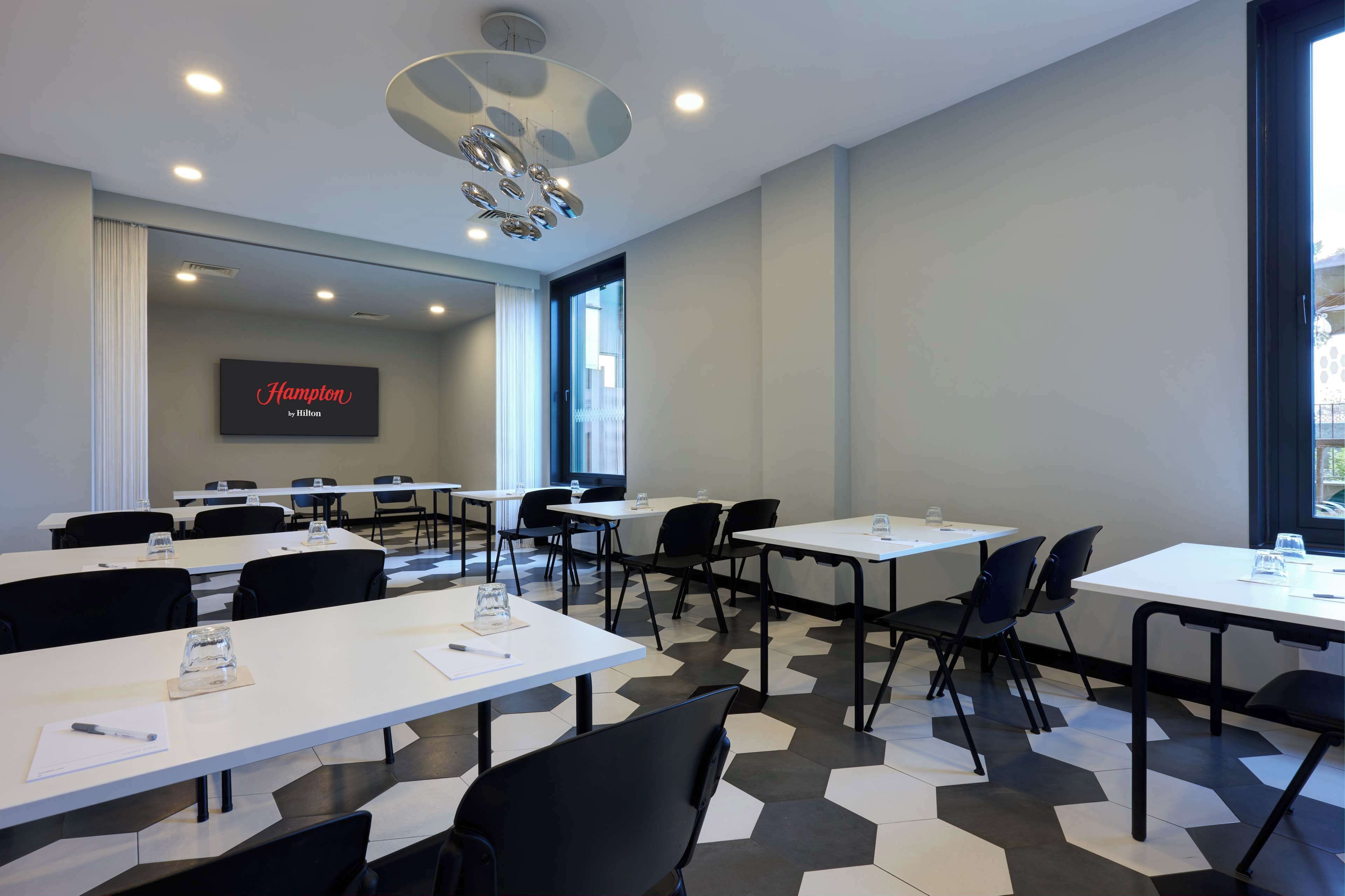 Ein Konferenzraum mit weißen Tischen, schwarzen Stühlen und einem großen Bildschirm, der "Hampton by Hilton" anzeigt.