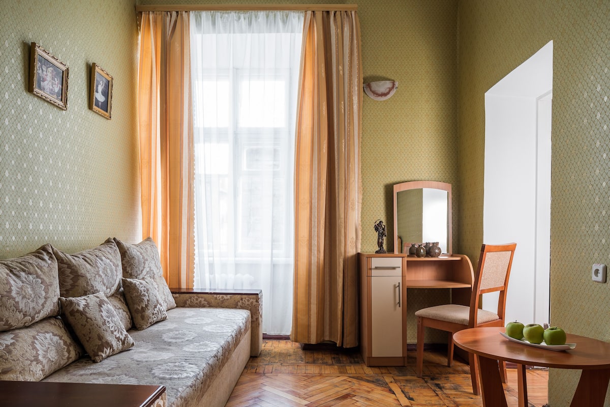 Una habitación con un sofá, una mesita con manzanas, una silla, un escritorio y un espejo.