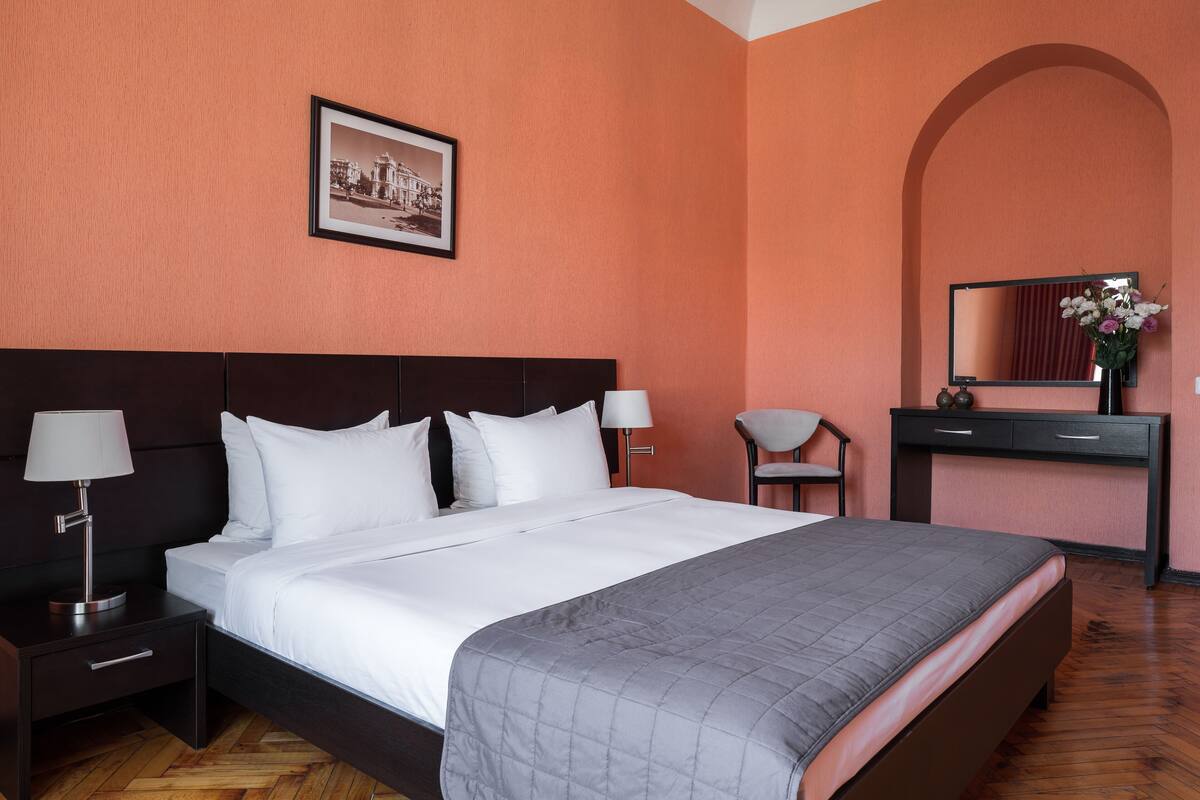 Una cama bien hecha con ropa blanca, un cabecero oscuro y una colcha gris acolchada. Hay una mesita de noche con una lámpara y un cajón, una pequeña mesa con una silla, y un escritorio con un espejo y un jarrón de flores.