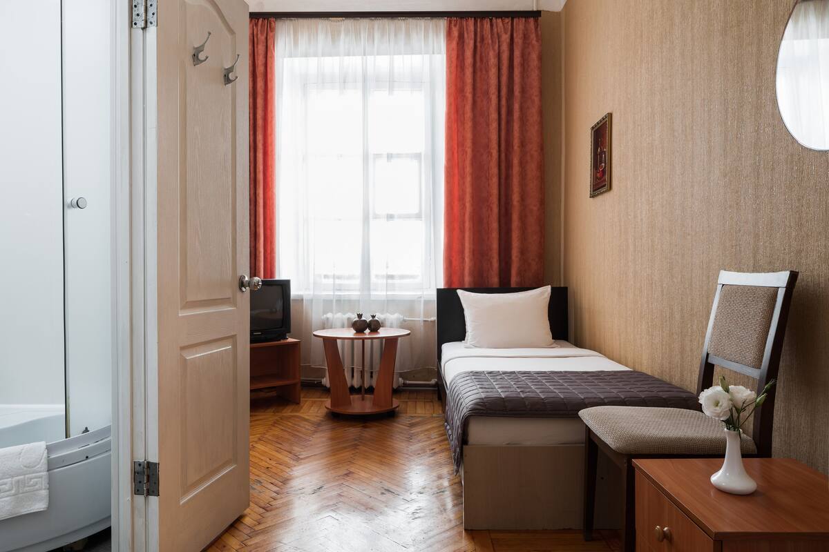 Una habitación de hotel compacta con cama, una mesita con un florero, una silla, televisión y ventana con cortinas.