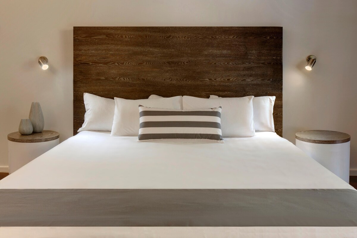 Una cama con cabecera de madera, ropa de cama blanca y dos mesitas de noche con lámparas.