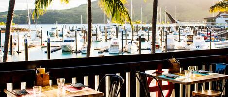 A restaurant with a view of a marina. Tables are set with menus and glasses.