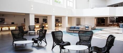 A spacious lobby with high ceilings, large windows, and a central seating area with black wicker chairs and tables.