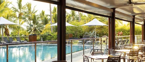 A poolside restaurant with tables and chairs, overlooking a pool and palm trees.