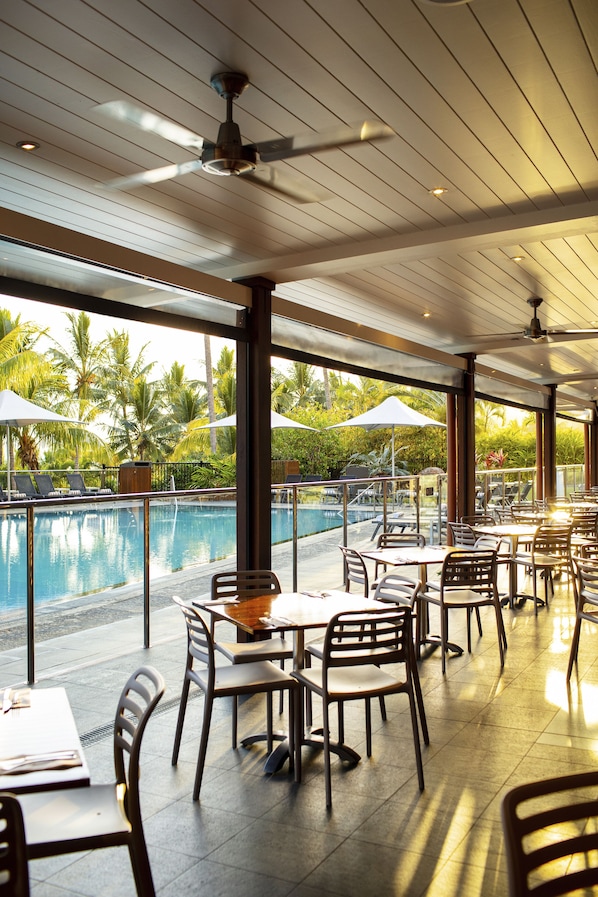Restaurante junto a la piscina con mesas y sillas, con vistas a una piscina y palmeras.