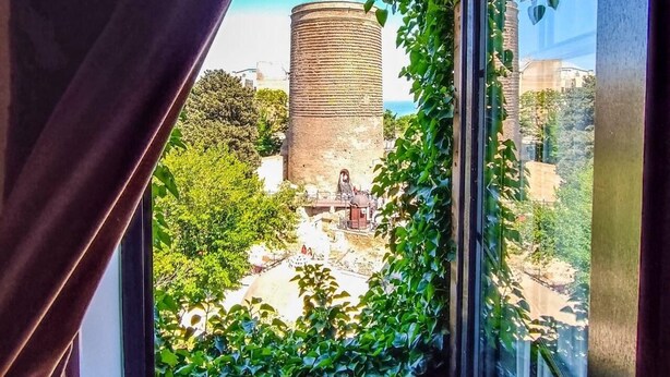 A view through a window with a curtain, featuring a cylindrical structure surrounded by greenery. - Sultan Inn Boutique (Baku)