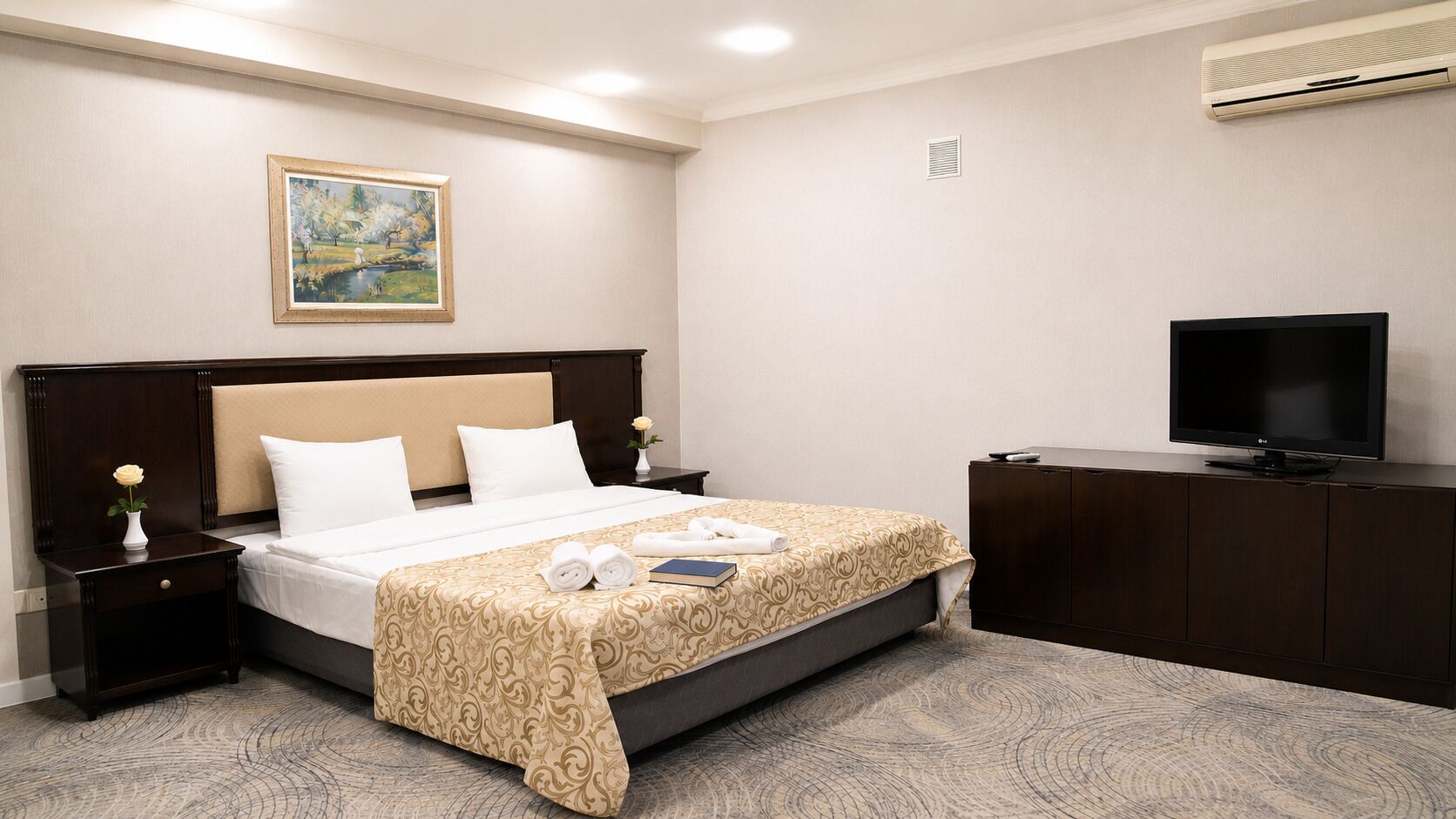 Una habitación de hotel con cama, televisor, un cuadro en la pared y una mesita de noche con un florero.