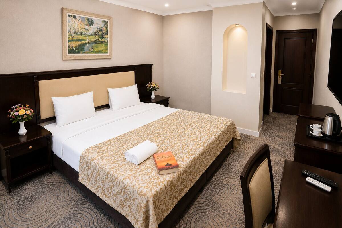 A hotel room with a large bed, a nightstand with a vase of flowers, a desk with a coffee maker, and a painting on the wall.
