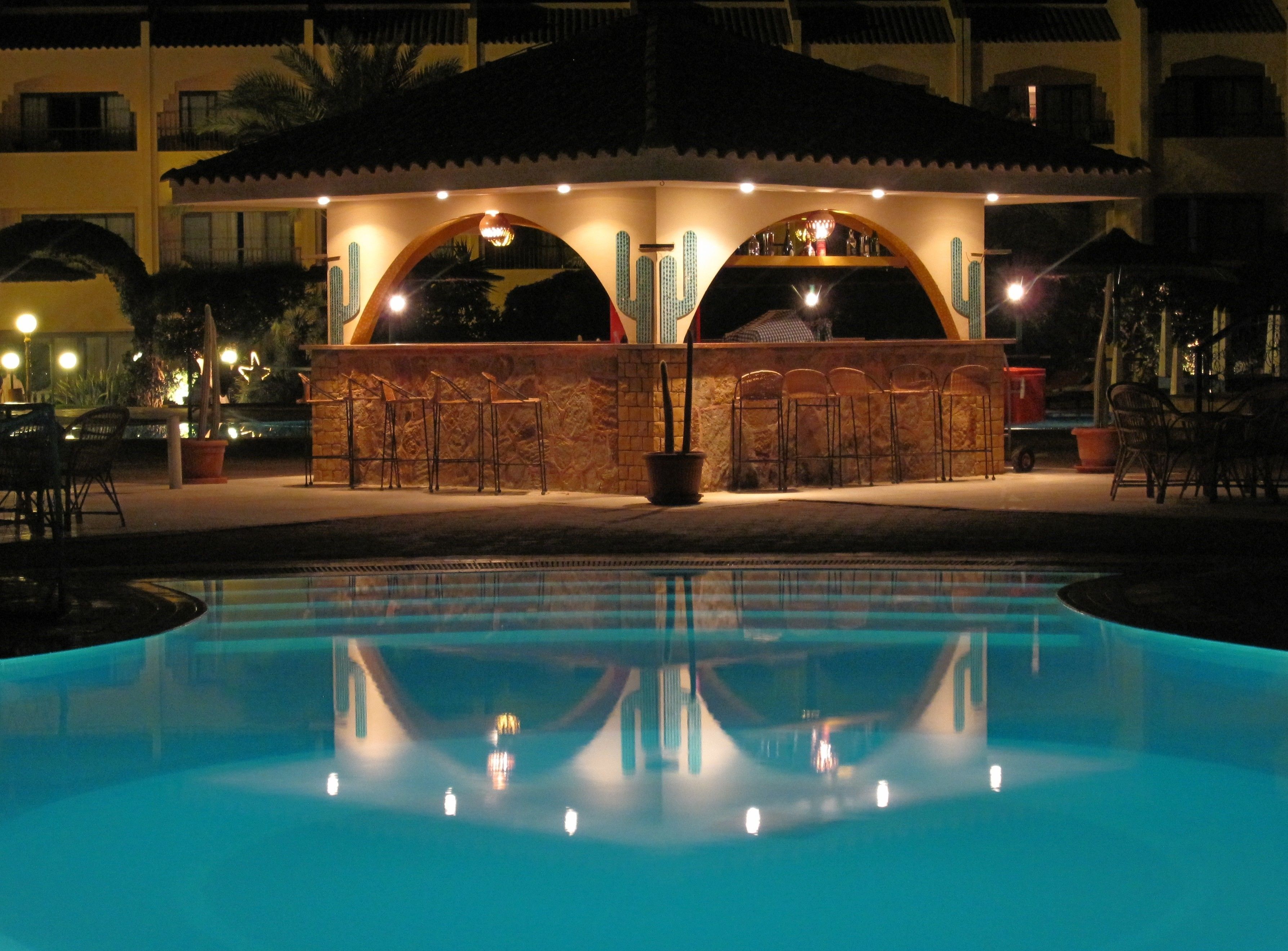 Un espace bar situé au bord de la piscine, avec la piscine en premier plan, entouré de chaises longues et d’un espace de détente couvert doté d’un toit en tuiles.